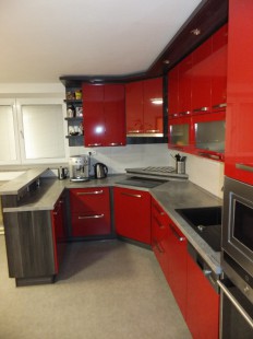 Kuchyň v odvážném červeném lesku, kombinovaného černošedým ořechem oddělující daný prostor menším barovým pultem.